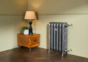 Ornate Rococo III cast iron radiator in Full Polish on feet with nickel valves, beside a marquetry side table and lamp on a herringbone floor.