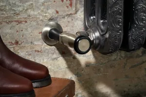 Close-up of an ornate Rococo III cast iron radiator in a dark reflective finish with a nickel angled valve, beside brown leather boots on a case.