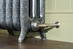 Close-up of an ornate Rococo III cast iron radiator in Full Polish on scrolled feet, with a satin nickel valve, green panelled wall and parquet floor.