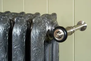 Close-up of ornate Rococo cast iron radiator columns in Full Polish with a nickel valve and black-trimmed head against green panelled wall.
