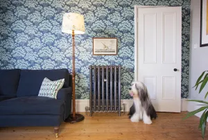Ornate Rococo III 970mm cast iron radiator in Satin Polish on feet against blue tree wallpaper, beside a navy sofa, floor lamp and a sheepdog.