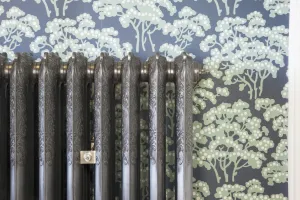 Close-up of an ornate Rococo III cast iron radiator in Satin Polish with a bleed valve, against blue wallpaper with pale green trees.