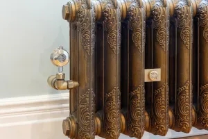 Close view of an ornate Rococo II 510mm cast iron radiator in Beaten Brass with a brass angled valve and wall stay, by a pale green wall and skirting.