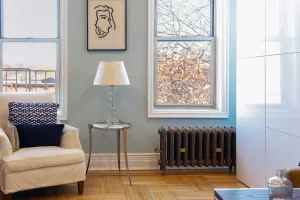 Ornate Rococo II cast iron radiator in Beaten Brass on feet beneath a sash window, beside a white armchair and side table lamp on parquet.