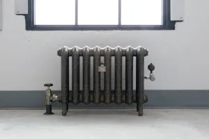 Ornate eight-section Rococo II cast iron radiator in Beaten Brass on feet below a window, with angled valves, on a concrete floor.