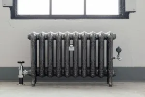 Ornate ten-column Rococo II cast iron radiator on feet in burnished silver-grey, with chrome valves, below a window on a concrete floor.