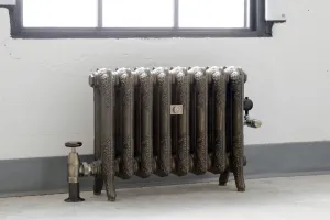 Ornate eight-column Rococo II cast iron radiator in Beaten Brass on feet beneath a window, with angled brass valves and a grey skirting.