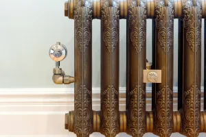 Close-up of an ornate Rococo II cast iron radiator in Beaten Brass with a brass thermostatic valve, against a pale wall and white skirting.