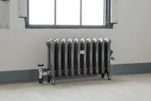 Ornate ten-section Rococo II cast iron radiator on feet in a silver-toned finish, with chrome valves, under a window on a concrete floor.