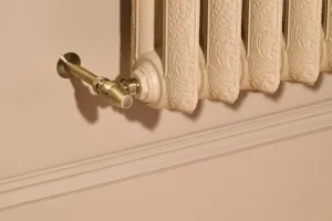 Close-up of an ornate Rococo II cast iron radiator in Farrow & Ball Planter No. G5 with a brass angled valve, against a pale pink wall with moulding.