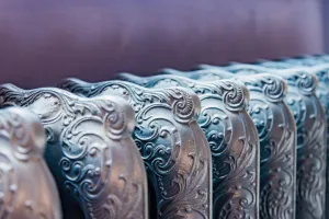 Close-up of the ornate scrolled sections of a Rococo III cast iron radiator in a silvery metallic finish against a purple wall.