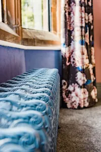 Close view of an ornate Rococo III 470mm cast iron radiator in pale blue beneath a stone window, beside floral curtains and grey carpet.