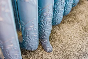 Close-up of the ornate scrolled columns and feet of a Rococo III cast iron radiator in a pale blue finish, standing on grey carpet.