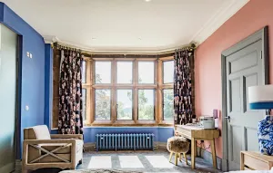 A low Rococo III cast iron radiator in pale blue beneath a mullioned bay window with floral curtains, in a bedroom with blue and pink walls.