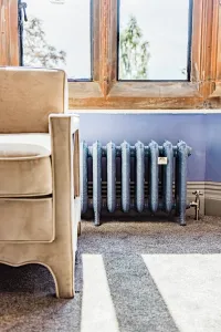 Ornate Rococo III 470mm cast iron radiator in blue-grey on feet beneath a stone window, with a brass valve, beige armchair and grey carpet.