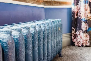Ornate Rococo III cast iron radiator in a mid-blue finish on feet, against a blue wall beside a floral curtain on grey carpet.