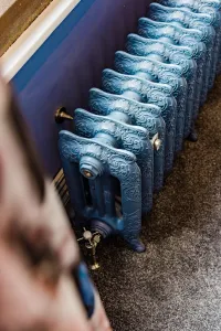 Ornate Rococo III 470mm cast iron radiator in a pale blue finish on feet, with brass valve and wall stay, against a navy wall on grey carpet.