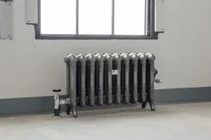 Ornate Rococo II 510mm cast iron radiator in Full Polish on feet below a window, with chrome valves, on a concrete floor.