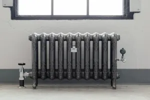 Ornate ten-column Rococo II cast iron radiator on feet in a dark metallic finish with polished detail, chrome valves, below a window.