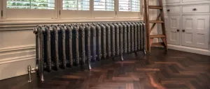 Ornate Rococo III cast iron radiator in Full Polish on feet beneath a shuttered window, with a valve, herringbone floor and a wooden ladder.