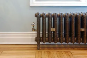 Ornate Rococo II cast iron radiator in Beaten Brass on feet below a window, with a chrome thermostatic valve, blue wall and parquet floor.