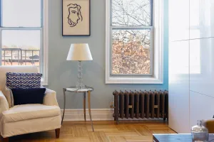 Ornate Rococo II 510mm cast iron radiator in Beaten Brass on feet beneath a sash window, beside a white armchair on a parquet floor.