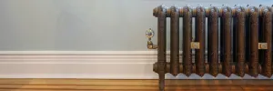Ornate Rococo II cast iron radiator in Beaten Brass on feet, with a brass valve and wall stays, against a pale blue wall above a wooden floor.