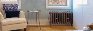 An ornate Rococo II cast iron radiator in Beaten Brass on feet beneath a window, beside a white armchair and glass side table on parquet.