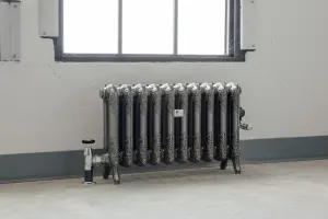 Ornate Rococo II cast iron radiator in Full Polish on feet below a window, with a chrome angled valve on a bare concrete floor.