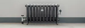 Ornate Rococo II 510mm cast iron radiator on feet, dark with polished highlights, chrome valves, against a grey wall on a concrete floor.