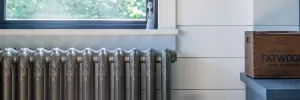Ornate Rococo II cast iron radiator in Grey Bronze below a window sill, against white panelled walls beside a wooden crate.