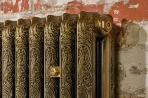 Close-up of an ornate Rococo II cast iron radiator in Aged Gold with swirling relief, a brass end plug and wall stay against a worn brick wall.
