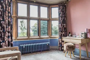 Ornate Rococo III 470mm cast iron radiator in blue on feet beneath a mullioned bay window with floral curtains, beside a wooden desk and stool.
