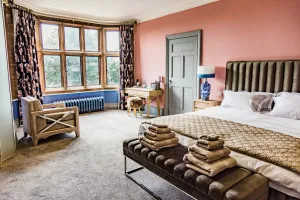 Ornate Rococo III 470mm cast iron radiator in pale blue, wall-mounted beneath a bay window in a pink bedroom with a large bed and armchair.