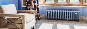 Low ornate Rococo III cast iron radiator in blue-grey on feet with valves, under a stone bay window beside a cream armchair.