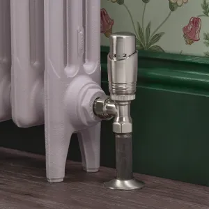 Niva angled thermostatic radiator valve in Satin Nickel on a pale lilac cast iron radiator, with green skirting and floral wallpaper behind.