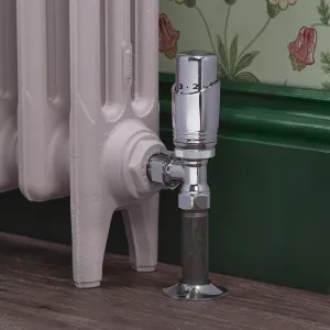 Chrome Niva angled thermostatic radiator valve on a pale pink cast iron radiator, beside a green skirting board and floral wallpaper.