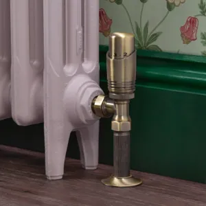Niva angled thermostatic valve in Antique Brass on a pale pink cast iron radiator, against a green skirting board and floral wallpaper.