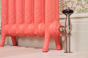 Windsor angled thermostatic valve in Satin Nickel with a black head, fitted to an ornate coral cast iron radiator on feet beside floral wallpaper.