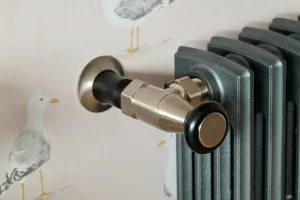 Close-up of a Windsor angled valve in brushed nickel with a black head, fitted to a dark grey Grace 4 Column radiator against seagull wallpaper.
