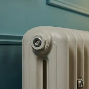 Close-up of a nickel bleed valve plug marked Castrads on the top of an Emmeline II radiator in Little Greene Portland Stone 77, against a teal wall.