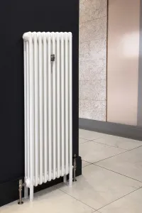 Tall white Florence 3 column steel radiator on feet against a navy wall, with satin nickel valves and a wall stay, on pale marble-effect tiles.