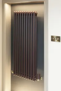 A tall wall-hung Florence 3 column steel radiator in a brown Cordoba finish with brass valves, set in a lit grey alcove beside a brass light switch.