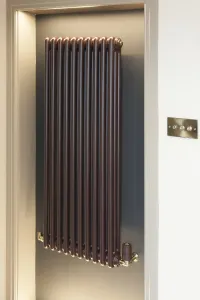 A tall wall-hung Florence 3 column steel radiator in a deep brown Cordoba finish with a brass thermostatic valve, set in a lit alcove.