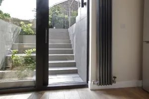 Tall vertical Florence 3 column steel radiator in dark grey beside a glazed door to tiled garden steps, with angled valves and oak floor.