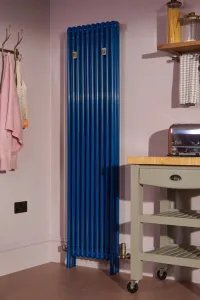 Tall Florence 3 Column 1800mm steel radiator in bright blue with brass Niva valves, in a pink kitchen beside a sage green trolley.