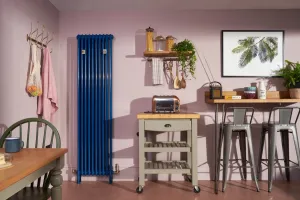Tall blue Florence 3 Column steel radiator, wall-hung with Niva valve, in a pink kitchen beside a butcher's block trolley and bar stools.