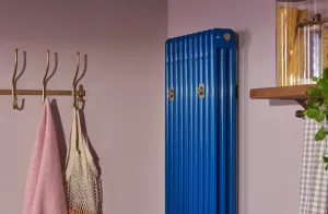 Tall Florence 3 Column steel radiator in gloss blue with brass wall stays, between brass coat hooks and a wooden shelf on a lilac wall.