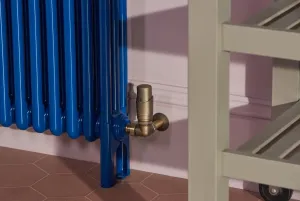 Base of a blue gloss Florence 3 column steel radiator on feet with a brass Niva thermostatic valve, pink wall and terracotta hexagonal tiles.