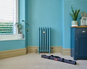 Emmeline III 870mm cast iron radiator in Bluegum on feet with brass valves, against a matching wall, with a grey tool bag on the carpet.